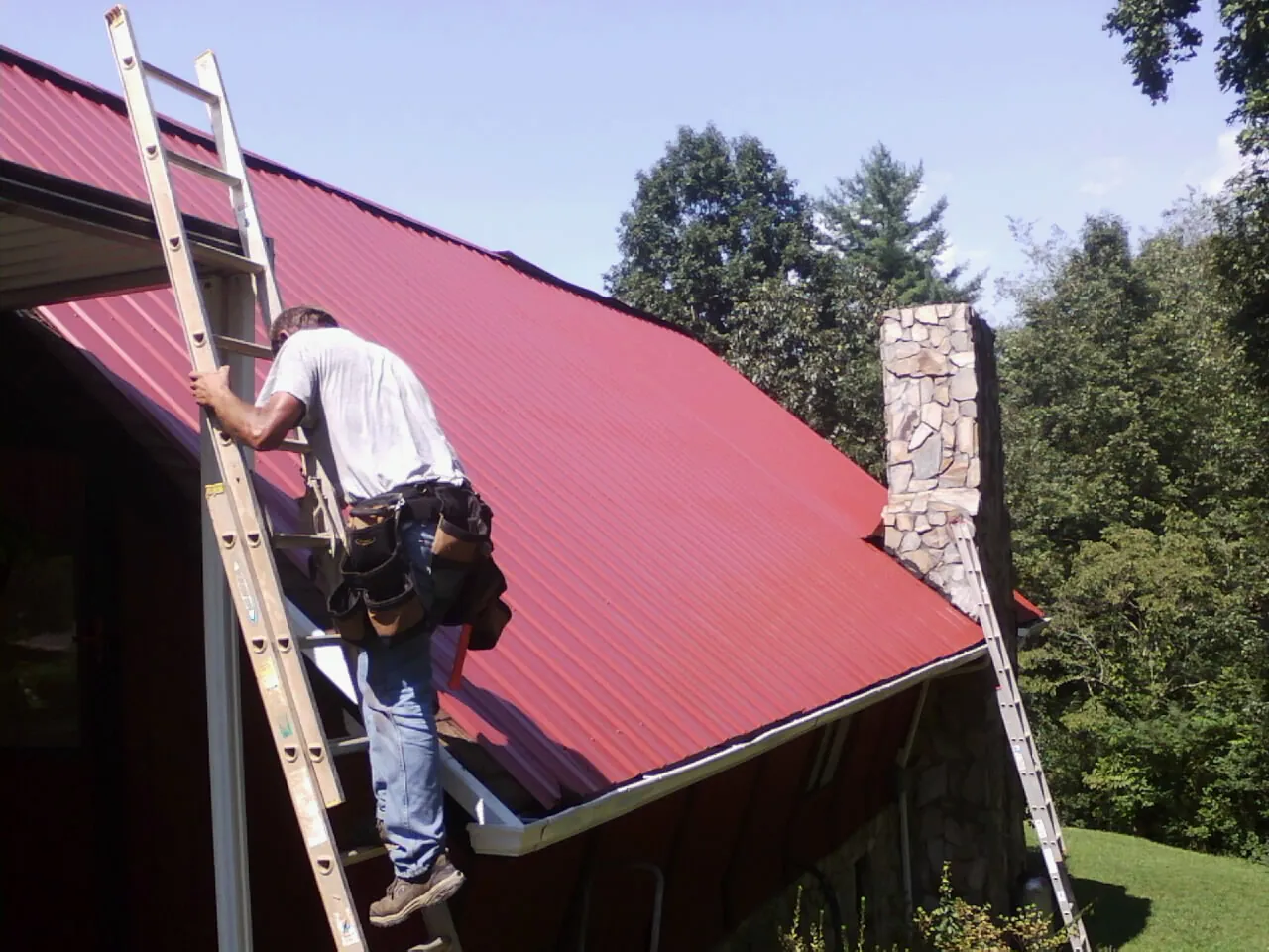 Roofer on a ladder beside a red metal roof and stone chimney — Soffit & Fascia Repair by Roofing Brothers in Denhoff
